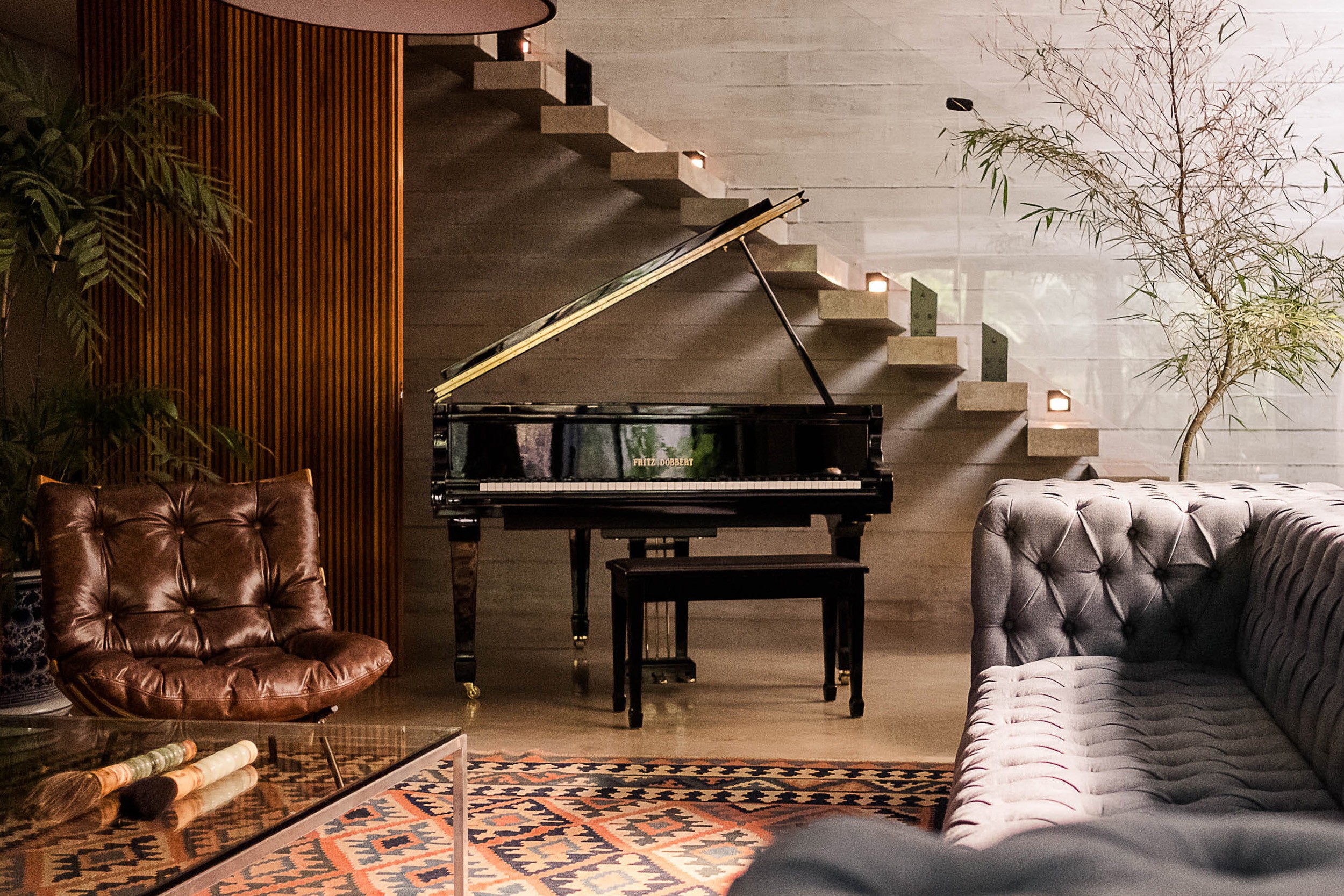 Sala elegante com piano de cauda preto em destaque ao centro. À esquerda, poltrona de couro marrom sobre painel de madeira. À direita, sofá Chesterfield cinza e, ao fundo, escada flutuante de concreto. Iluminação suave e natural entra pela lateral, destacando um tapete estampado no chão.