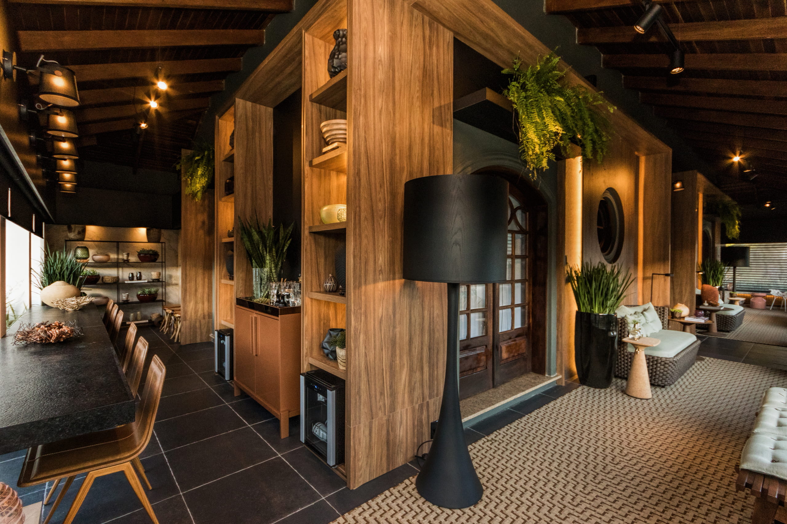 Vista interna de ambiente sofisticado com teto de madeira. À esquerda, há um balcão com banquetas. No centro, uma divisória de madeira com prateleiras iluminadas e uma luminária de piso preta em primeiro plano. À direita, área de estar aconchegante com tapete, sofá e plantas sob iluminação quente indireta.