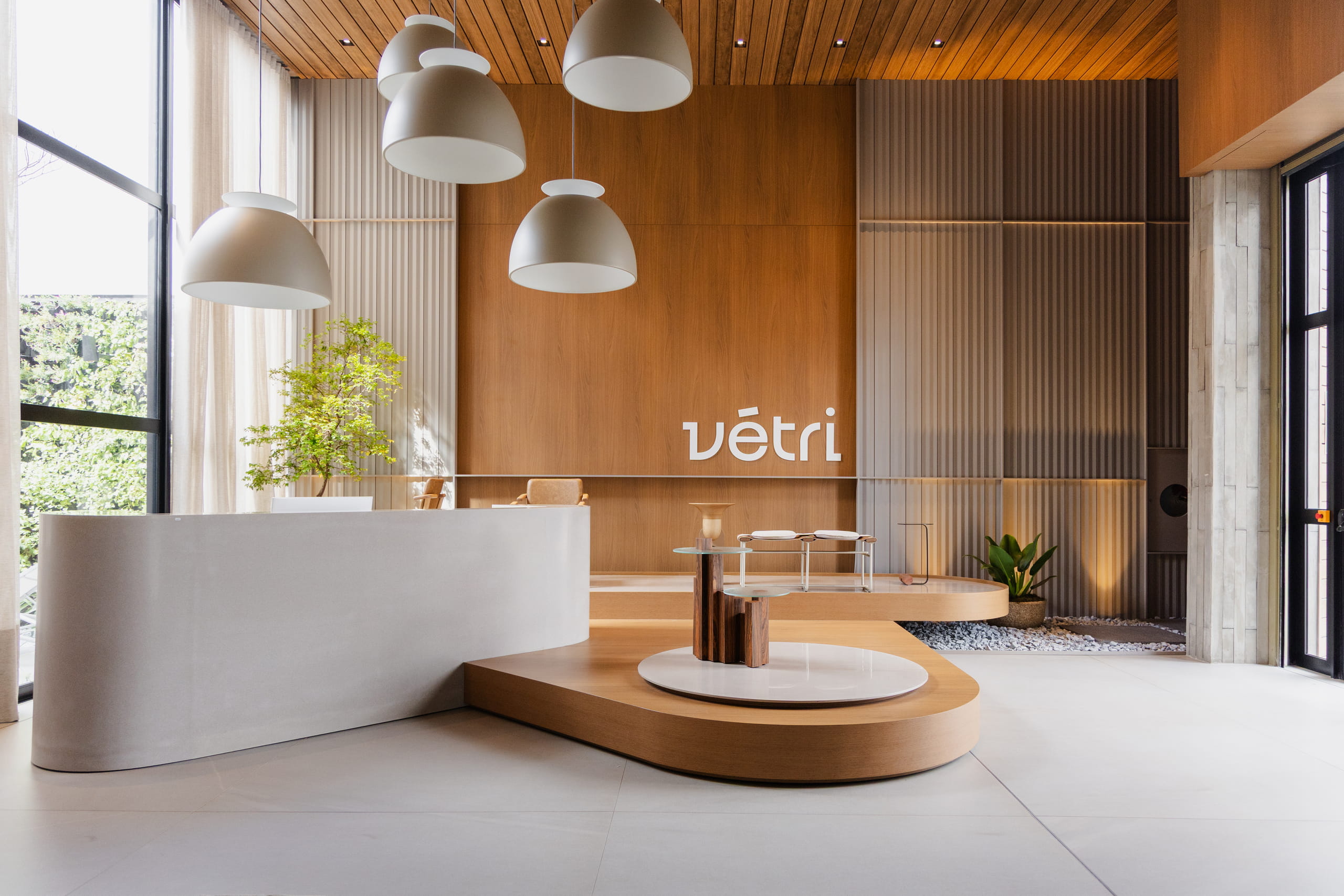Uma foto de uma recepção de hotel de design contemporâneo. No primeiro plano à esquerda, há uma mesa de recepção branca e curva. Atrás dela, uma parede de madeira clara com o logotipo "vétri" em branco. Pendurados no teto, quatro lustres em forma de domo de cor bege. Do lado direito, painéis acústicos texturizados e, no chão, uma área elevada com uma mesa lateral de vidro e plantas. O teto é forrado com ripas de madeira, criando um ambiente sofisticado e convidativo.