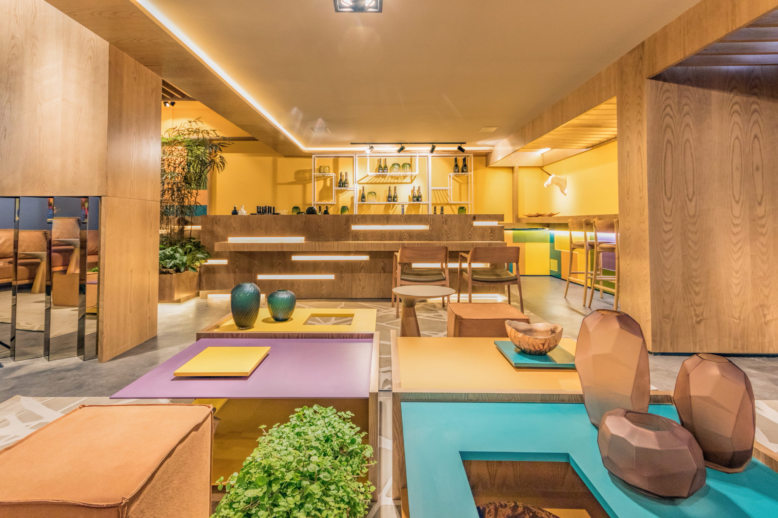 Interior moderno com painéis de madeira clara, paredes amarelas e piso de cimento cinza. O teto tem iluminação de LED. No primeiro plano, móveis baixos de madeira com superfícies coloridas (roxo, turquesa, laranja), decorados com vasos de cerâmica e plantas. Ao fundo, um balcão de madeira escalonado com luzes de LED e prateleiras com garrafas de vinho. Cadeiras de madeira e estofamento estão próximas. Plantas em vasos ladeiam o espaço.