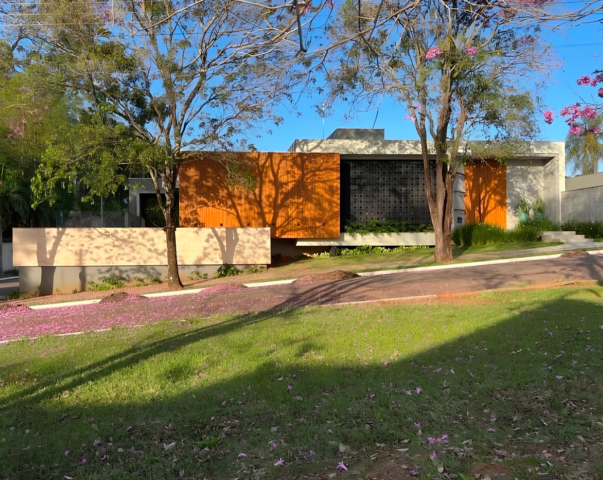 Fachada de uma casa moderna de um pavimento sob céu azul. A construção combina painéis alaranjados, detalhes em concreto aparente e um cobogó escuro no centro. Em frente, há árvores com flores cor-de-rosa, um gramado verde e a calçada salpicada de pétalas rosa caindo das árvores. Uma mureta bege baixa acompanha a calçada à esquerda.
