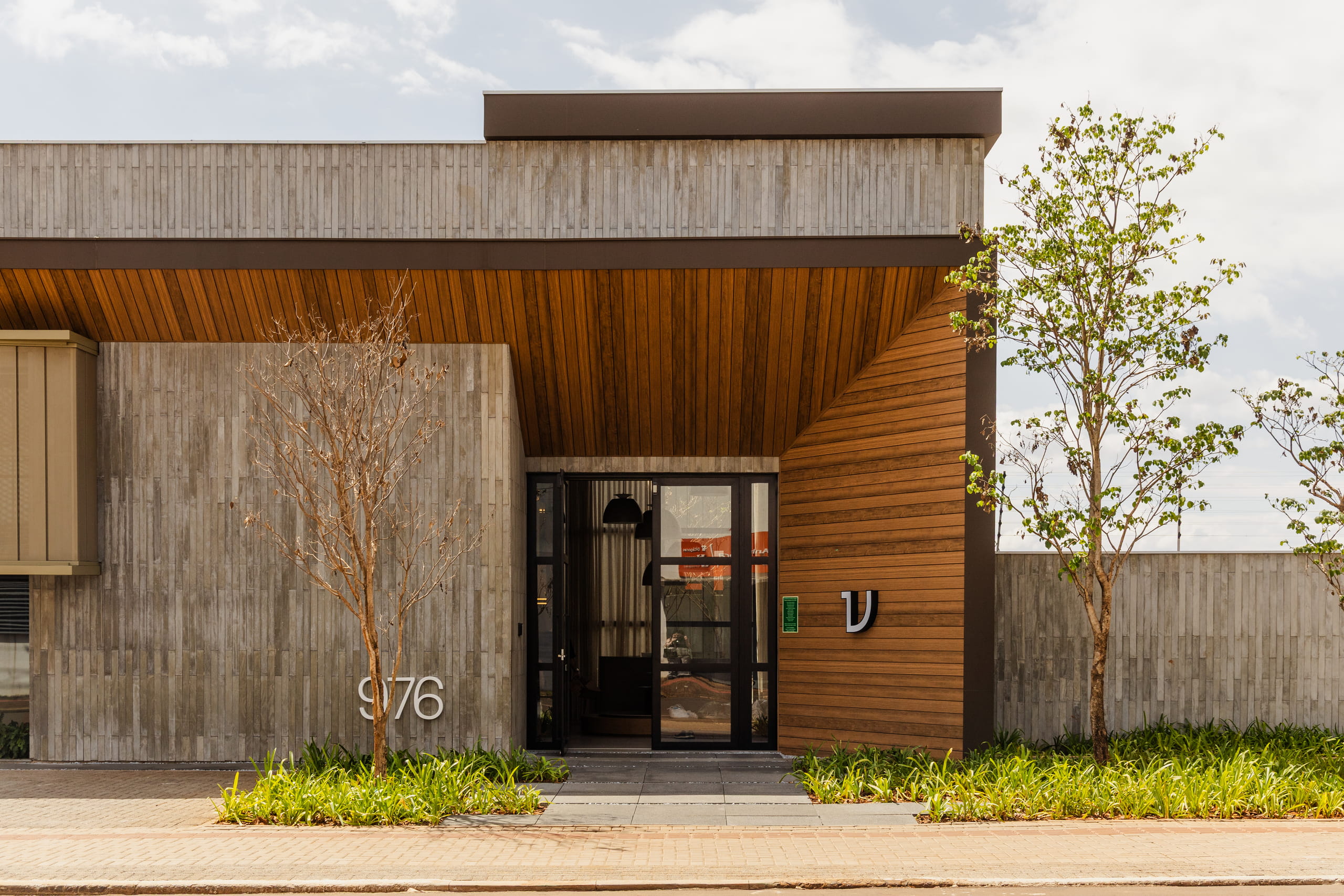 Fachada moderna de arquitetura contemporânea com paredes em concreto aparente e entrada com forro em réguas de madeira. Porta principal de vidro com esquadria preta. À esquerda, o número "976" afixado na parede, acompanhado por pequenas árvores e vegetação rasteira na calçada. À direita, um logotipo prateado decorativo na parede de madeira.