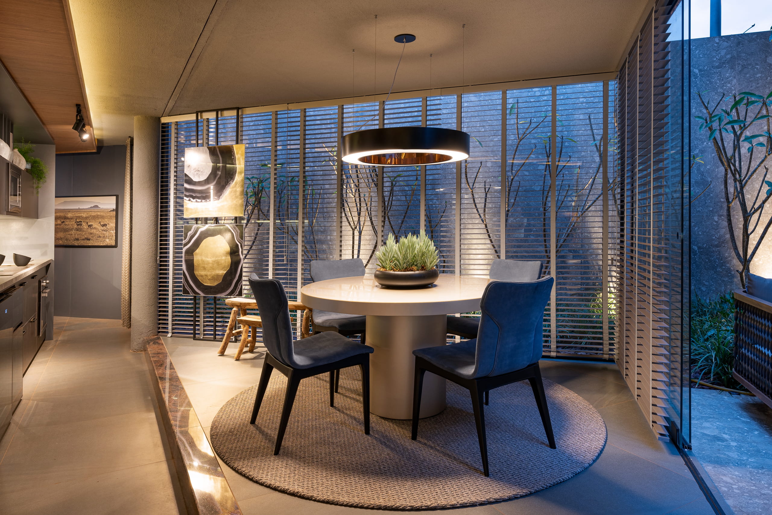Modern dining room with a beige round table on a circular rug, surrounded by four dark blue upholstered chairs. A central vase and black ring pendant light complete the setting, while large windows with horizontal blinds reveal soft lighting and plants in an indoor garden.