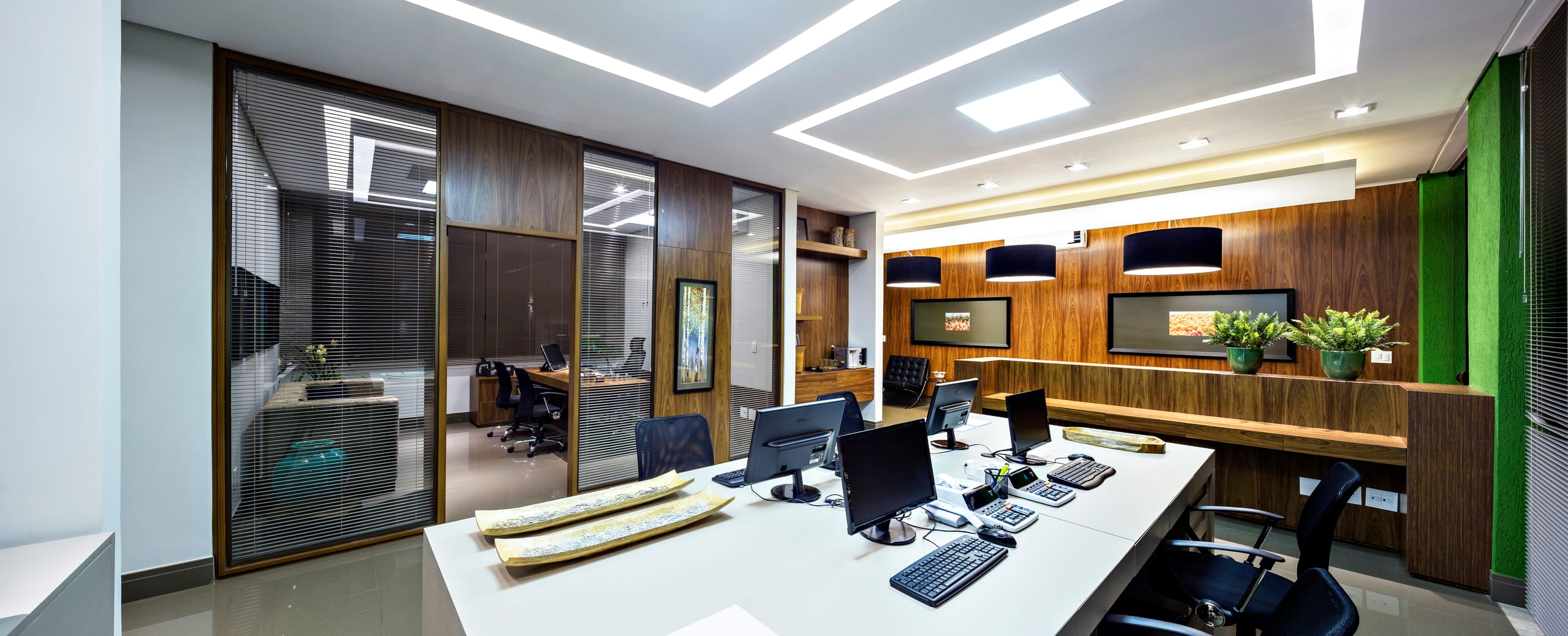 Escritório corporativo moderno com grandes bancadas de trabalho brancas equipadas com computadores e cadeiras pretas. À esquerda, divisórias de vidro e madeira delimitam salas privadas. Ao fundo, uma parede com painéis de madeira, luminárias pendentes pretas e vasos de plantas sobre um móvel planejado. O teto possui iluminação LED retangular embutida.