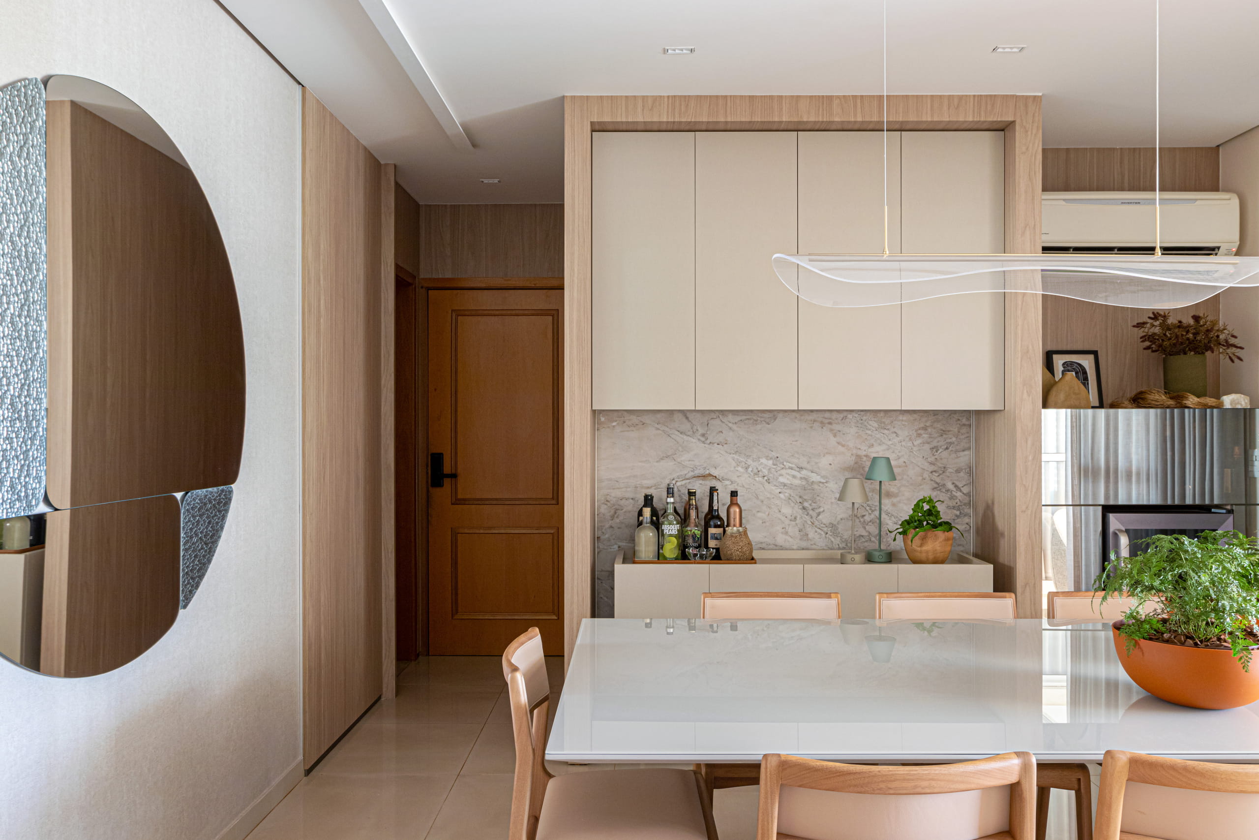 Sala de jantar integrada e moderna. Em primeiro plano, mesa branca com cadeiras de madeira e vaso terracota com planta. À esquerda, grande espelho orgânico na parede clara. Ao fundo, buffet com cantinho do bar, armários aéreos beges moldados em madeira, painel de mármore e luminária pendente translúcida e ondulada sobre a mesa. Ao fundo à direita, geladeira de inox.